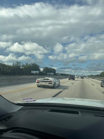 The image captures a highway scene in Boca Raton, United States, observed from the interior of a vehicle. In the midground, a white Lamborghini Diablo, displaying the license plate "VNJSZ", travels in the second lane from the left. Further ahead in the same lane, a dark-colored Jeep Wrangler is visible. Additional vehicles, including cars and a truck, are present in the distant lanes. The highway features concrete median barriers to the left and clearly marked yellow lane lines. In the background, dense green foliage lines the highway's periphery. Two billboards are positioned above the left-side foliage, one partially visible with the text "LOTUS". The sky above is blue with numerous scattered white cumulus clouds. A dark dashboard and a purple sticker are partially visible in the immediate foreground of the image.