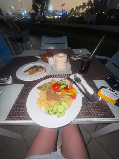 The image shows a table with a place setting at what appears to be an outdoor restaurant in Unknown, Egypt. On the table are a glass of dark liquid with a straw, cutlery, a plate with pasta, salad and meat, and another plate with a fish dish. A cylindrical object wrapped in what looks like fabric sits on a small dish next to a brown object. A person's leg and part of their clothing are visible to the right of the table. The background shows what appears to be a city scene with lights and palm trees.