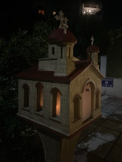 The image depicts a miniature replica of a church or bell tower, possibly a decorative element or a scaled-down architectural model. It is made of a light-colored brick-like material with red roof sections and a red dome. The structure has arched windows, some of which are illuminated from within, suggesting a light source inside. There are also decorative elements resembling crosses on top of the structure. The miniature is situated outdoors, surrounded by lush green foliage from trees and bushes, and appears to be at night, as indicated by the darkness and artificial lighting. In the background, a street sign with Greek lettering is visible, reading "ΠΕΝΙΖΕΛΟΥ 4 Β-Α" which translates to "Penizelou 4 B-A". This suggests the location is on Penizelou street in Greece, specifically in Neoi Epivates based on the provided context. The overall impression is a quaint, nighttime scene focusing on the miniature church structure.