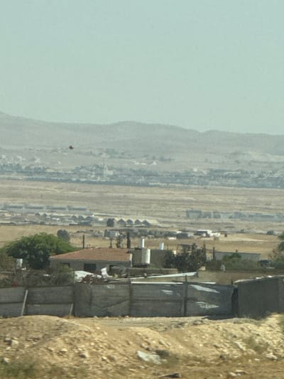A wide shot of a cityscape in the Al-Qasoum Regional Council, Israel, with a mountain range in the background on the left side. A red object floats in the distance over the mountain range. The ground is beige and brown. Buildings are scattered in the center of the image with what appears to be a wall on the right side of the image. A tree is visible in front of the wall.