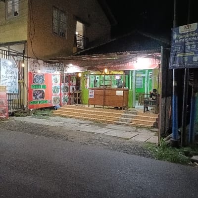 The image shows a small restaurant in the town of Kaliwungu, Indonesia, located at the coordinates -6.961, 110.251. The restaurant is located on a street corner and has a canopy over an outdoor seating area with a few tables and chairs. The restaurant is illuminated with strings of lights. The menu board advertises a variety of dishes, including "Mie Cak" and "Mie All." A lone customer sits at a table. An adjacent building, painted yellow, is visible to the left. There is a large billboard on the side of the building on the right. The scene is lit by streetlights and appears to be captured after dusk. The street is paved, and there are small plants and shrubs along the sidewalk.