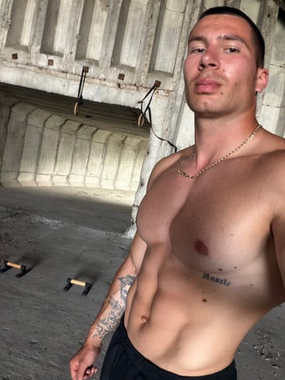 A shirtless male individual is positioned in the foreground, capturing a selfie within a raw, unfinished concrete structure in Izbeglii, Bulgaria. The subject has a muscular physique, short dark hair, and a neutral expression, looking directly into the camera. He wears a thin gold chain necklace around his neck and dark athletic shorts on his lower body. Distinct tattoos are visible: the word "Hustle" in an Old English font on his left rib cage, and a detailed black ink design covering his entire left forearm, including what appears to be a figure or face near the elbow.

The background consists of textured, grey concrete walls and a ribbed concrete ceiling. Two sets of wooden gymnastic rings with black straps are suspended from anchors embedded in the ceiling. On the uneven, dusty concrete floor, two pairs of black push-up bars with light brown handles are present. The overall lighting suggests natural light entering the space, casting subtle shadows.