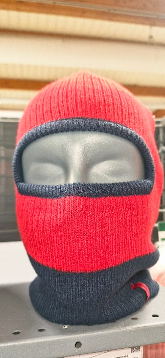 A red and dark blue knitted balaclava is displayed on a dark gray, semi-gloss mannequin head in Vendin-le-Vieil, France. The balaclava's upper section, including the crown, is red, transitioning to a dark blue portion around the lower neck and chin. A dark blue knitted band outlines a single, large oval opening designed for the face. A small red fabric loop is visible on the lower right side of the dark blue section. The mannequin head rests on a gray, perforated metal shelf. The blurred background features dark, possibly gridded structures directly behind the subject, topped by horizontal light-colored panels.