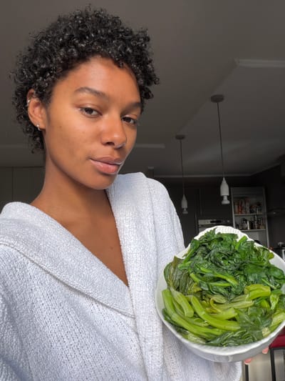 A person with curly black hair, wearing a white robe, is holding a white bowl of cooked leafy green vegetables. The background includes a kitchen cabinet and pendant lights. The location cannot be determined from the image.