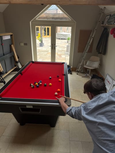 The image depicts a man playing pool in a room located in Sand Hutton, United Kingdom, at the coordinates 54.025 latitude and -0.933 longitude. The man is positioned at the end of a pool table with a red felt surface. He is holding a pool cue in his right hand, preparing to strike a white cue ball that rests on the table. There are multiple multi-colored pool balls scattered across the surface, some grouped together and some isolated. The room features a large, arched window with double glass doors leading to an outdoor patio. The patio is visible through the glass doors. The patio is paved with a patterned stone design and lined with shrubs. There are two metal ladders leaning against the wall, a white board hanging on the wall and a dark green coat hanging on a hook. The room has a wooden ceiling, and the walls are painted white.