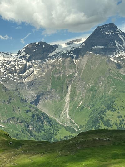 This is a vertical, medium-shot photograph of the Grossglockner mountain in Fusch an der Großglocknerstraße, Austria. The mountain is immense, with a dark, rocky peak partially covered in snow. The slopes of the mountain are a mix of steep, gray rock faces and lush green grass, with patches of snow scattered across the upper reaches. A deep gully runs down the mountainside, appearing to be a dry riverbed or a path carved by erosion. The lower slopes are covered in dense green forestation. In the foreground, rolling green hills are visible, dotted with small, winding paths. A tiny, dark-colored wooden structure, resembling a small hut or shed, is nestled on the left side of the foreground hills. The sky above is a vibrant blue, with large, fluffy white clouds drifting across. The lighting suggests it is daytime, with the sun casting shadows that highlight the textures and contours of the mountainous landscape. There are no people or animals visible in the image.