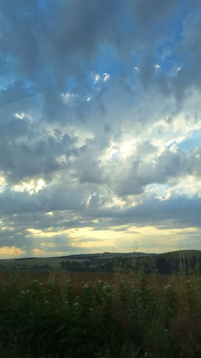 The image captures a wide-angle view of a landscape dominated by a dramatic sky over rolling hills. The sky is filled with a mix of dark, moody clouds and patches of bright, ethereal sunlight breaking through. The sunlight casts visible rays down towards the horizon. Below the sky, a layered landscape unfolds. In the foreground, tall grasses and wildflowers create a textured, natural border. Beyond this, fields of varying colors, suggesting crops or wild vegetation, stretch towards a distant treeline and the gentle slopes of hills. The overall impression is one of a natural, serene environment under a dynamic and partly overcast sky, possibly indicative of approaching or receding weather. The specific location in Radomirtsi, Bulgaria, is not visually discernible from the image itself.