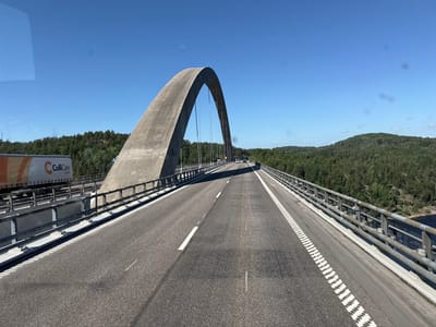 A wide, gray, asphalt road stretches across a modern concrete bridge, curving slightly to the right. The bridge is characterized by a prominent, tall concrete arch that rises majestically on the left side, with thin cables extending from its apex to support the roadway. The sky above is a clear, vibrant blue, suggesting a bright, sunny day. Dense green forests cover the rolling hills that flank the bridge, with glimpses of a dark body of water visible on the right side, hinting at a natural landscape.

On the left side of the bridge, a white semi-trailer truck with an orange and white logo that reads "ColliCare Logistics" is visible. The truck appears to be traveling in the same direction as the viewer. Further down the road, a few other vehicles can be seen, also moving away from the camera. The road itself is marked with white lines, indicating traffic lanes. The metal railings along the sides of the bridge are a weathered gray.

The scene conveys a sense of travel and transit through a scenic, possibly rural or semi-rural, environment. The clear sky and bright sunlight indicate daytime, likely mid-morning or afternoon. The overall impression is one of a functional, modern infrastructure integrated into a natural setting.