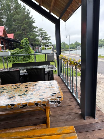 The image depicts an outdoor seating area of a restaurant or cafe, likely located in Belebei, Russia, given the context provided. The foreground features a wooden picnic table with a mosaic-tiled tabletop, accompanied by a wooden bench. Behind this, a covered patio area is visible, furnished with several black wicker chairs around glass-topped tables. A grey railing with decorative spindles separates the patio from a paved walkway and a body of water, possibly a pond or lake, with fountains in the distance. The surrounding environment includes lush green trees and foliage, a red-roofed building, and overcast skies. The overall scene suggests a tranquil, perhaps slightly damp, outdoor dining experience.