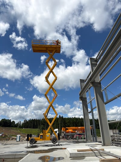 A construction site is depicted under a partly cloudy blue sky. In the foreground, a yellow scissor lift is extended, with one worker wearing a safety harness and orange vest at the top platform. To the right, the steel framework of a building is under construction, with large grey beams. In the middle ground, a portable toilet, a white truck, and an orange crane are visible. The background features a dense forest. The location is identified as Janowice, Poland.