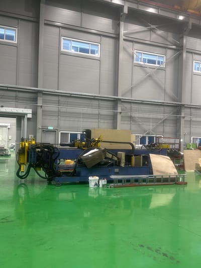 The image depicts a large industrial workshop in Pyeongtaek-si, South Korea, characterized by its high ceilings and gray, corrugated metal walls. Prominently featured is a large, blue and yellow industrial machine, likely a pipe bending machine, situated in the center of the frame. The machine has numerous hoses, levers, and a long metal rod extending from it. In front of the machine, on the highly reflective green floor, are a silver ladder and several white buckets. The floor shows some smudges and a reflection of the machinery and lighting. To the left, a large roll-up door is partially open, revealing more of the workshop's interior. Windows with white frames are visible along the upper walls. The overall environment suggests an active manufacturing or fabrication facility. There are no people visible in the image.