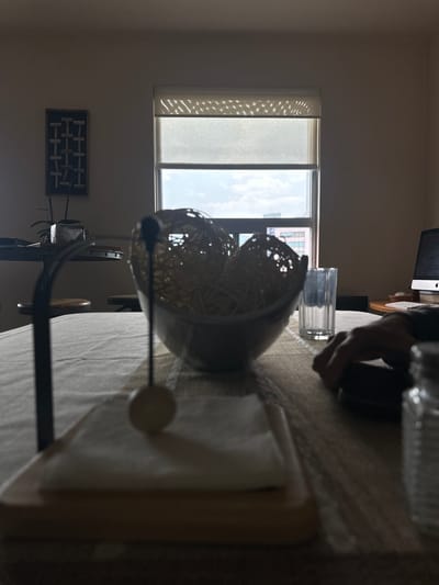 The image shows a dimly lit interior scene. A window with partially drawn blinds is visible on the left. A table is covered with a textured tablecloth and holds several items: a bowl, a drinking glass, and a stack of napkins in a napkin holder. A computer monitor is visible in the background. It is not possible to determine the location or any specific details related to Jesús del Monte, Mexico from the image alone. There are no people visible, and no actions or interactions are depicted.