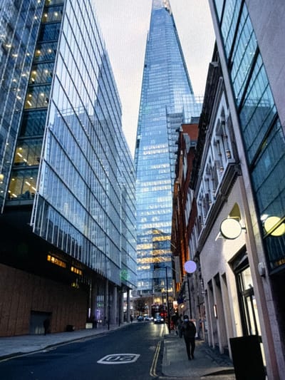 This image captures a street scene in London, United Kingdom, with the distinctive Shard skyscraper prominently featured in the background. The perspective looks down a street flanked by buildings.
On the left, a modern, multi-story building with a facade primarily composed of blue-tinted glass and metal framework rises sharply. Its numerous windows reflect the overcast sky and show interior lights illuminated in a warm yellow hue. The lower section of this building features a darker, possibly stone or metal, facade. A faint human figure is visible near the base of this structure. The asphalt road in the foreground displays a white "20" speed limit marking, indicating a 20 mph zone, and double yellow lines are painted along the left curb.
On the right, a series of buildings of varying architectural styles line the street. The closest building has a lighter-colored facade with glass windows and two circular, externally illuminated signs – one white and one yellowish-green. Further down, buildings with older, reddish-brown brickwork and ornate white window surrounds or balustrades are visible. A single individual, dressed in dark clothing and wearing a face mask, walks away from the camera on the paved sidewalk on the right. Another purple circular sign is affixed to a building facade further along this side.
In the distance, towering over the urban landscape, is The Shard, a tall, glass-clad pyramidal skyscraper with a distinct tapered top. Its reflective surface and interior lights are visible, indicating its presence as a major landmark. Behind the foreground structures, streetlights, additional pedestrians, and vehicles, including a red double-decker bus, are discernible, suggesting a busy urban environment. The sky is uniformly bright and overcast, creating diffused light, but the activation of artificial lights in buildings and signs suggests either early morning or late afternoon/evening.
