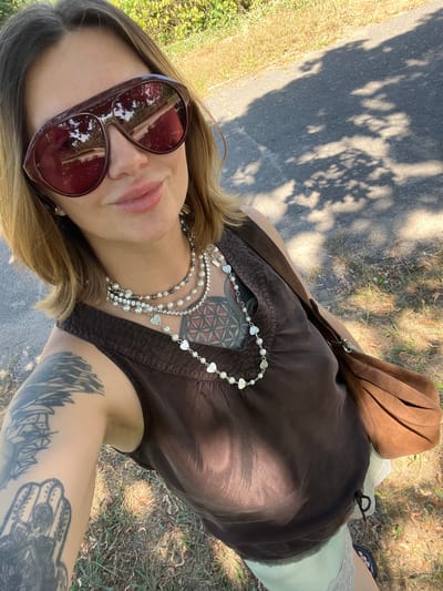 A woman takes a selfie outdoors on a sunny day. She is wearing large, dark red sunglasses, a brown sleeveless top, and a light-colored skirt with lace trim. She has multiple necklaces, including pearl and heart-shaped pendants. She has tattoos on her left arm. The background shows a paved path, grass, and trees. The location is specified as Voznesensk, Ukraine.