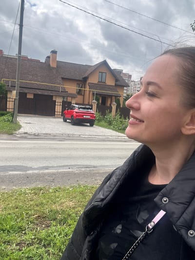 A medium shot captures a woman in a black puffer jacket looking to her right with a slight smile. She has brown hair pulled back and is wearing a black t-shirt with a subtle butterfly print. In the background, a red SUV is parked in the driveway of a two-story brick house with a dark-tiled roof. The driveway is paved with interlocking grey stones. A light pole stands to the left of the house. The scene is outdoors with a cloudy sky overhead. The location is the Medvedevo Urban Settlement, Russia.