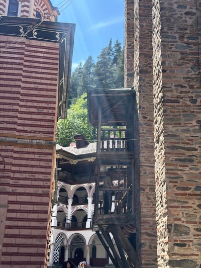 The image depicts the exterior of a building in Rilski manastir, Bulgaria, featuring stone masonry, red-tiled roofs, and an ornate arcade with black-and-white checkered detailing. A wooden structure stands between the stone wall and the building, and a tree is visible in the background. There are individuals present in the arcade. The image is taken from a low angle looking upwards.