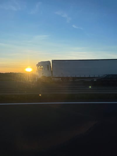 The image depicts a highway scene at sunrise or sunset near La Palme, France. A large white semi-trailer truck, consisting of a cab and a light grey box trailer, occupies the central portion of the frame, traveling from left to right. The sun, a brilliant yellow-orange orb, is low on the left horizon, partially obscured by the truck's cab, generating significant lens flare and atmospheric haze. A dark-colored passenger car is visible in the lane behind the truck's trailer. The immediate foreground shows a dark asphalt road surface with a solid white line, indicating a multi-lane highway viewed from a moving vehicle. The sky transitions from bright orange and yellow near the horizon to clear light blue at the top. A dark, indistinct landmass or vegetation line is silhouetted against the horizon. No specific individuals are clearly discernible within the vehicles.