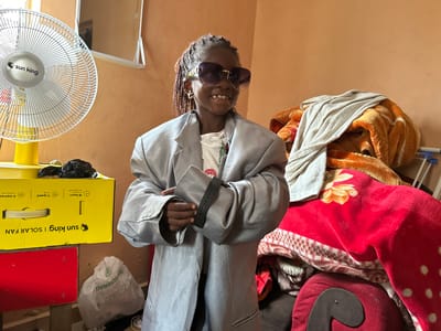 In a room with peach-colored walls in Gidan Waziri, Nigeria, a young person with braided hair and large sunglasses stands with their arms crossed. They are wearing an oversized gray blazer over a white t-shirt. The person is also wearing hoop earrings and a dark smartwatch. To the left of the person, a yellow box with "Sun King | Solar Fan" printed on it sits on a red surface. A white fan with the "Sun King" logo is to the left of the box. To the right of the person, a pile of blankets and what appears to be a red cushion are visible. A white plastic bag with "Reusdblog" printed on it rests on the floor near the red surface.