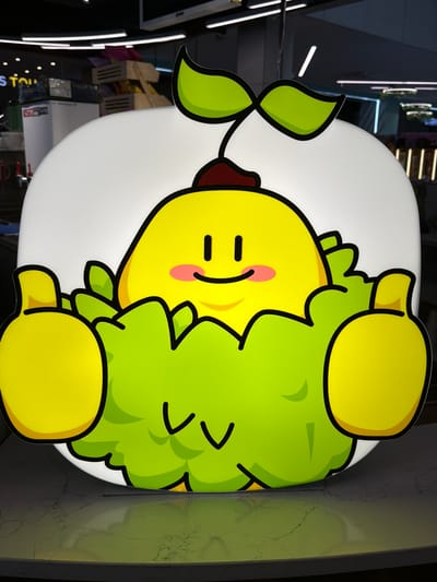 A brightly lit, cartoonish sign of a yellow, round character with green leafy accents and large yellow hands giving a thumbs-up is displayed in what appears to be a retail or commercial establishment. The character has a simple face with black dots for eyes, a curved black line for a mouth, and two pink circles for cheeks, suggesting a cheerful expression. It is wearing a small, dark red cap. The sign is backlit by a white glow. The background is blurred, showing out-of-focus elements of shelves and lighting fixtures, indicative of an indoor environment. The sign is placed on a reflective, marbled surface. The overall impression is that of a whimsical advertisement or decorative element. Based on the provided information, this scene is located in Ulaanbaatar, Mongolia.