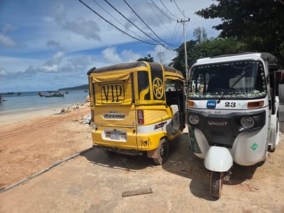 Two tuk-tuks are parked on a dirt road next to a sandy beach with calm ocean waters. The tuk-tuk on the left is yellow with black trim and features the word "VIP" prominently on its side, along with a star logo and honeycomb pattern. It also has "BAJAJ Maxima" branding and a license plate reading "1L-8184" and "PREAH SHANOUK". A spider graphic is visible on the rear of this vehicle.
The tuk-tuk on the right is white and also displays "Bajaj Maxima" branding. It has a license plate-like sticker with "ABA" and the number "23" on the front. The windshield is decorated with small shells. An "LPG" sticker is visible on the front of this tuk-tuk.
In the background, several fishing boats are moored in the shallow water, and a lush green, tree-covered hill or island is visible in the distance. The sky is partly cloudy with a bright blue expanse. The lighting suggests it is daytime, likely late morning or early afternoon, and the weather appears warm and sunny. The overall scene evokes a tropical, coastal atmosphere.