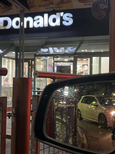 The image shows a McDonald's restaurant at night in Gelemenovo, Bulgaria. The iconic "McDonald's" sign is prominently displayed in white lettering against a dark background. In the foreground, a car's side mirror reflects a white car driving through a drive-thru lane. The drive-thru area appears to be equipped with metal poles and barriers. Inside the restaurant, through the large glass windows, a few people are visible, and brightly lit menu boards display food items. The overall scene suggests a typical evening operation at a fast-food establishment.