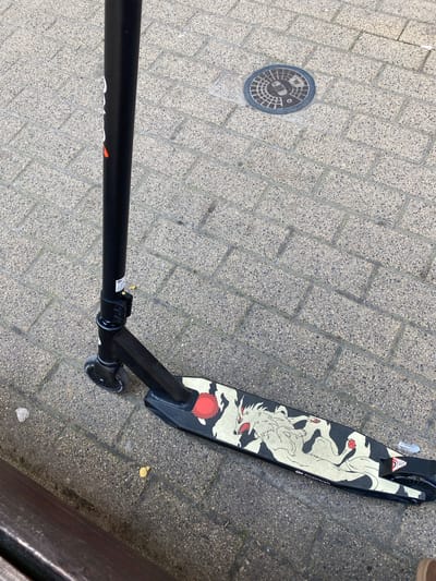 A black kick scooter with a stylized white wolf graphic on its deck is lying on a brick-paved sidewalk. The scooter is angled diagonally across the frame, with its handle pointing upwards and to the left, and its deck extending towards the bottom right. A round metal manhole cover is visible on the sidewalk to the upper right of the scooter. The sidewalk is made of light-colored bricks arranged in a herringbone pattern. The image is taken from a high angle, looking down.