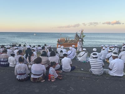 In the city of Abang, Indonesia, a group of people are gathered on a beach, facing the ocean. The sky is a soft gradient of blue and yellow, indicating either sunrise or sunset. The ocean is calm, with a few small boats visible in the distance. In the foreground, the beach is covered in dark sand.

The people are seated on the ground, many wearing traditional Balinese attire. Some are dressed in white, with head coverings, suggesting they might be participating in a religious ceremony. In the center of the gathering, a decorated altar or offering table is set up, adorned with fruits, flowers, and figurines, with a person in white standing next to it.

The crowd consists of both adults and children, with women wearing lace tops and patterned sarongs, and men in white shirts and pants or traditional patterned wraps. A young girl in a colorful outfit is sitting closer to the water, playing in the sand. The overall atmosphere appears to be one of reverence and communal gathering.