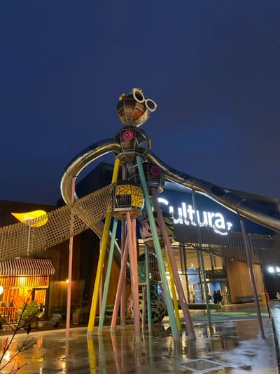 A whimsical, metal sculpture of a figure wearing sunglasses stands outside the "Cultura" building. The figure is composed of interconnected, basket-like spheres connected by silver tubing. Brightly colored rods extend from the figure's body, appearing to penetrate the glass wall of the building. The surrounding ground is wet, reflecting the lights of the building. The sky is a deep blue, suggesting it is night.