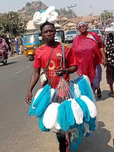 A man in a red soccer jersey stands on a street carrying a large bundle of blue and white netting. A red-dressed woman stands nearby. He has some netting piled on his head. In the background, there is a yellow auto-rickshaw and various buildings and a hill. The netting is mostly white and blue, but some of the netting has silver.