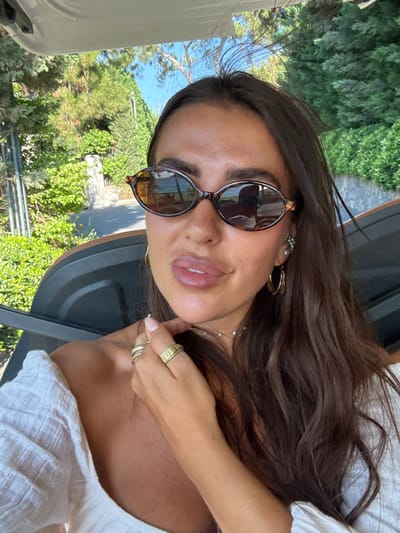 A woman is taking a selfie in a golf cart in Bodrum, Turkey. She has long, dark brown hair and is wearing sunglasses and a white off-the-shoulder top. She is also wearing several rings and earrings. The background is a lush green landscape with trees and a blue sky. The sun is shining brightly.