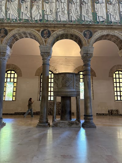 A photograph, rotated 90 degrees counterclockwise, shows an interior space, likely a church or basilica, in Ravenna, Italy. The composition is dominated by ancient architectural elements, including columns, arches, and a pulpit or ambo.

On the left side of the image is a mosaic wall featuring rows of figures in white robes. The arches above are decorated with intricate patterns and circular portraits.

Two prominent columns, made of marble, are visible in the foreground. They support an archway and an elaborately carved pulpit or ambo. The pulpit is adorned with geometric designs and symbols.

Arched windows, arranged in rows, are visible in the background. Light streams through the windows, creating bright beams on the floor.

A woman stands in the foreground. She is holding a mobile phone.

The floor is made of marble tiles.