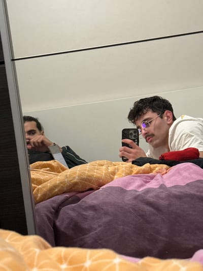 A person with curly hair and glasses poses for a selfie in a mirror. They hold a phone with a patterned case. The reflection shows a person with darker hair. A white t-shirt is visible on the selfie-taker. In the foreground, colorful bedding in shades of purple, pink, and yellow obscures part of the view. The background appears to be a white wall or cabinet. The overall scene seems casual and domestic.