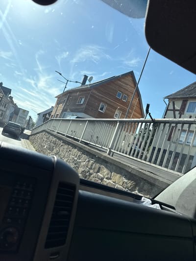 This media file captures a view from inside a vehicle, looking outwards through the windshield and side window. The scene depicts a street in what appears to be a European village, with traditional German architecture visible.
The primary subjects are buildings, including a prominent wooden house with several windows and a white-paneled structure behind a metal railing. The buildings have a rustic, aged appearance, suggesting an older settlement. A stone wall lines the side of the road, adding to the historical feel of the location.
In the foreground, part of the vehicle's interior is visible, including a black dashboard with an air vent and control buttons. A white crumpled object, possibly a tissue, is tucked into a compartment.
The activity taking place is implied by the perspective from within a vehicle – likely driving through or parked in the village. The sky is a clear blue with wispy white clouds, indicating daytime and fair weather. The sun is high in the sky, casting shadows and creating a bright, clear scene.
Notable details include the specific architectural styles of the houses, the stone wall, and the presence of a satellite dish on top of one of the buildings. A street lamp is also visible. In the background, other vehicles, including a truck with visible text, are present on the road. The text on the truck appears to be in German, with phrases like "Wir bewegen was" and "Wir packen das."
The overall impression is of a peaceful, sunny day in a picturesque German village. There are no visible people, and the emotions conveyed are neutral, focused on the observational nature of the image. The setting is clearly urban or semi-urban, with residential buildings and a street.