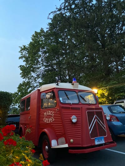 A red, vintage Citroën H Van food truck, identified by "MARC'S CREPES" lettering on its side and a "CREPE" Connecticut license plate, is centrally positioned in a paved parking area during twilight in Ridgefield, United States. The vehicle features a cream-colored roof, a blue beacon light, and an additional spotlight mounted on the roof. Interior lights illuminate the vehicle's windows. To the right and rear of the red truck, a blue hatchback is partially visible. To the left and rear, a dark SUV is partially visible. Red geraniums are in bloom in the immediate foreground, and large, dark green trees fill the background against a pale blue and light-hued sky. A bright yellow street lamp is illuminated on the right side of the frame, casting light on the scene.