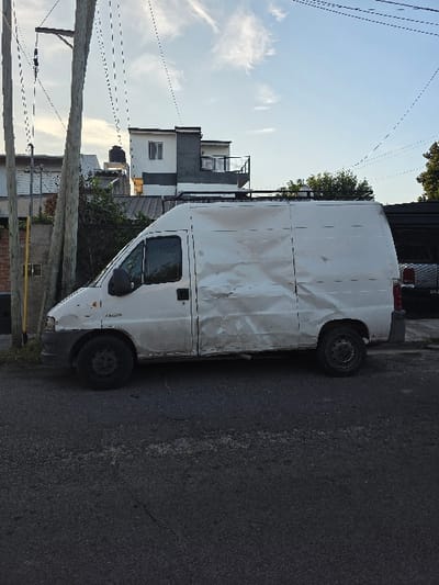 A white cargo van, with visible dents and damage along its side panels, is parked on an asphalt road. The vehicle has a roof rack, and its tires appear worn. Adjacent to the road, there's a telephone pole and partial views of buildings with various architectural styles, some featuring balconies. Overhead, power lines stretch across the sky.