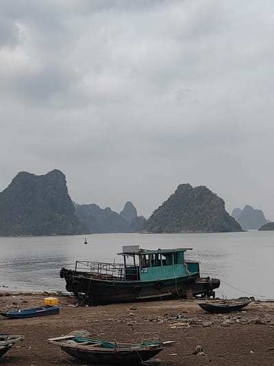 The photo depicts a seascape scene in Hải Phòng, Vietnam. The scene is overcast with a grey sky. The sea stretches to the horizon, with several karst islands rising from the water. In the foreground, boats rest on the shore. One is a larger, green-painted vessel. Smaller boats are scattered around. Debris litters the beach.