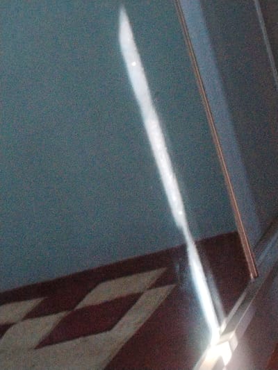 The image depicts an interior scene with a dark blue wall and a tiled floor. A diagonal band of bright light, possibly from a window or a light fixture, cuts across the wall and floor, creating a reflective effect on the glossy floor. The floor has a patterned design of alternating dark red and light-colored diamond shapes. A metal railing is visible on the right side of the image. There are no people present in the image. The scene appears to be a hallway or a room with a polished floor. The location in the city of Kulob, Tajikistan is not ascertainable from the visual information provided in the image.