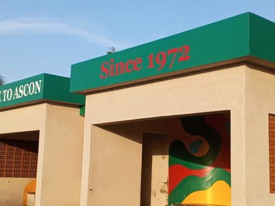 The image depicts two buildings at the entrance of ASCON in Tawpo, Nigeria. The building on the left reads "Welcome to ASCON" in white lettering on a green rectangular sign. The building on the right has a similar green sign that reads "Since 1972" in red lettering. Both buildings are beige in color and have open archways. The archway on the left building leads to a patterned brick wall, while the archway on the right building reveals a wall with an abstract, colorful mural featuring red, green, yellow, and blue paint. The sky above is blue with some visible clouds.