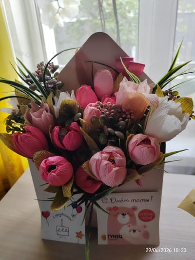 An envelope-shaped bouquet of handcrafted paper flowers, predominantly in shades of pink and white, is presented on a wooden table. The bouquet is arranged in a light brown envelope holder that opens at the top. The flowers, resembling tulips and other floral shapes, are intricately folded from textured paper. Some greenery and smaller, dark-colored berry-like elements are interspersed among the flowers.
On the front of the envelope holder, there are two distinct decorative panels. The left panel features a simple line drawing of a red balloon, a birthday cake with a lit candle, and a star, along with the Russian phrase "С праздником!" (Happy Holiday!). The right panel displays a cheerful design with two cartoon bears, one larger pink bear and a smaller white bear cub. The larger bear has a red circular badge on its chest with the Russian text "ЛЮБЛЮ ТЕБЯ!" (I Love You!). Below the bears, there is a logo with the Russian word "ТУНЕЦ" and smaller text.
In the background, a window with white sheer curtains is visible, allowing soft, diffused light to enter the scene. The curtains are slightly yellowed at the edges, possibly due to sunlight. The overall environment appears to be an indoor setting, likely a home or a gift shop. The image also has a timestamp in the lower right corner: "29/06/2026 10:23". The location is identified as Maladzyechna, Belarus.