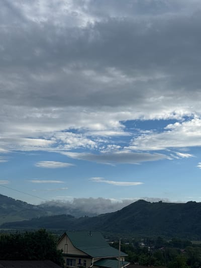The image captures a panoramic view of a mountainous landscape under a cloudy sky, likely taken from an elevated position within or near Soloneshnoye, Russia. In the foreground, the steeply pitched, dark green tiled roof of a house is prominently visible, with a lighter colored facade and windows. To the left, the dark green foliage of trees partially obscures the view.
The middle ground reveals more houses with similarly colored roofs and the dark green, dense forest covering the lower slopes of rolling hills and mountains. Power lines crisscross the scene, adding a man-made element to the natural setting.
The background is dominated by a range of lush, dark green mountains, their peaks softened by patches of mist and clouds clinging to their upper reaches. The sky above is a dramatic mix of dark, heavy grey clouds in the upper portion and lighter, scattered white clouds against patches of bright blue in the lower half, suggesting a dynamic weather system. The overall impression is one of a rural, mountainous area with scattered human settlements amidst a vast natural landscape.