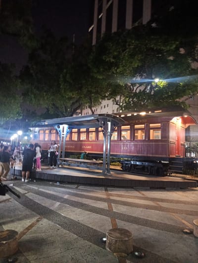 The image depicts a night scene in the city of Guayaquil, Ecuador, at a historical railway exhibit. A restored red train car, numbered 115, is prominently displayed on a platform. The car has large windows that are illuminated from within, suggesting it is a heritage exhibit. The text "PRIMERA CLASE" is visible on the side of the train car, indicating it was once a first-class carriage. A covered waiting area with benches is situated next to the train. Several people are gathered around the exhibit, some standing on the platform and others in the foreground, observing the train. The ground in front of the platform is paved with a patterned surface. Trees and a tall building are visible in the background, illuminated by streetlights, contributing to the urban night ambiance.