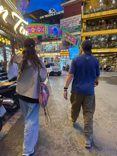 The image depicts a street scene in Sa Pa Ward, Vietnam, featuring two individuals, one male and one female. The male is wearing a blue shirt, grey pants, and a dark-colored cap, seen walking in the foreground. The female, seen on a motorcycle, is wearing a light-colored jacket, blue jeans, and a dark cap, with a pink and floral backpack.
The scene is illuminated with colorful, decorative lights and signs, including "Sapa Trekking & Souvenirs," "Anh Đào Spa," "Đồ Chơi - Quà Tặng," and "Hô Restaurant." A car is parked in the background. The street pavement is visible, and buildings are lined up on both sides of the street, providing a vibrant atmosphere.
