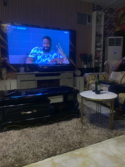 This image depicts an interior living room scene located in Akpajo, Nigeria. A large, dark-framed flat-screen television is centrally positioned, displaying a show from "SHOWCASE" with a "16L" rating. The screen shows a bearded man wearing a patterned blue and white shirt, with subtitles reading "You should be scared."

Below the television is a dark-colored entertainment unit featuring both open and closed storage, including light-colored drawers. In the foreground, a black, ornate coffee table with gold detailing and a reflective surface is visible. To the right, a round side table with a white marble top and gold-colored metal legs holds a dark green wine bottle. A person is seated on a cream and gold upholstered sofa to the right, with only their lower legs, clad in dark trousers, and a wrist wearing a watch visible.

The floor is covered partially by a shaggy, light grey area rug, transitioning to light-colored tiled flooring towards the bottom right. The walls display distinct finishes: the left wall features vertical wooden paneling with a window-mounted air conditioning unit near the ceiling, while the right wall is adorned with a patterned wallpaper. A tall, white shelving unit with decorative items, including artificial flowers in vases, stands against the patterned wall, next to a floor-standing air conditioning unit displaying "17". Additional decorative elements include red artificial flowers, a framed picture, and a vintage-style clock with Roman numerals on a shelf near the sofa.