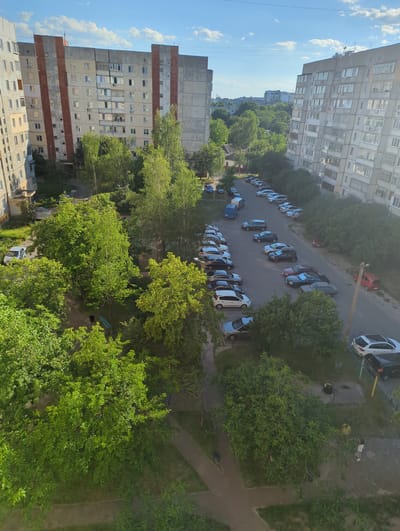 This media file captures an aerial view of a residential area in Boryspil, Ukraine, during daylight hours under a clear, blue sky with scattered clouds. The scene features a street lined with parked cars, interspersed with mature green trees. Apartment buildings, typical of Soviet-era architecture, dominate the background, with their multi-story facades and numerous windows visible.
The primary subjects are the cars parked along the road and on a dirt patch. Several people are visible in the frame; one person in a yellow shirt is standing near a tree, and another, wearing light-colored clothing, is walking. A few cars are in motion or have their doors open, suggesting normal activity within the neighborhood. The abundance of trees indicates a park-like or green urban setting. The lighting suggests it is a sunny day, likely late morning or afternoon. The overall impression is a typical, everyday scene in a suburban or urban residential area. No visible text is present in the image.