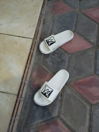 The image captures two white slide sandals resting on a tiled surface. Each sandal features a broad white strap adorned with a distinctive black and white logo, which appears to be a stylized "W" or "MW" within a black square frame. On the outer side of the strap, near the sole, there is small, barely legible text in black, possibly indicating a brand name like "VILLANUEVA."
The setting is a transitional area between two different types of flooring. On the left, a section of large, light-colored (beige or cream) square tiles with dark grout lines is visible, suggesting an indoor or covered space. This area is bordered by a raised, dark brown strip, possibly wooden or a decorative tile, which separates it from the other surface. On the right, the sandals rest on an outdoor-style pavement composed of hexagonal interlocking stones. These stones form a repeating geometric pattern of dark gray/black and reddish-brown rhombuses.
The scene is lit by bright, even daylight, though no specific time of day or weather conditions are explicitly indicated. There are no individuals present, and no particular activity is occurring; the sandals are simply placed on the ground. The context provided specifies the location as Kecamatan Panji, Indonesia.