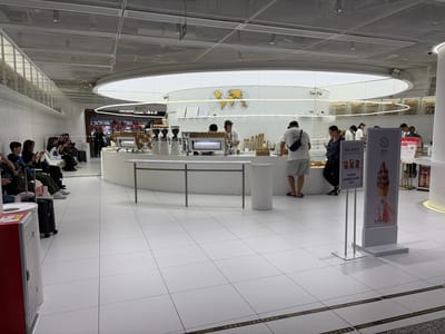 The image shows the interior of an % Arabica coffee shop, likely located in Baiyun District, China. The scene is a modern, minimalist setting with white tiled floors and a circular coffee counter. A world map graphic is displayed on the back wall above the counter. Customers and baristas are present. Some customers are seated, waiting with luggage, while others are standing near the counter. A display sign advertises "White Americano" with a picture of an ice cream cone. Signs with both English and Chinese text are visible. Markings on the floor read "Keep Clear Under Fire Protection Shutter".