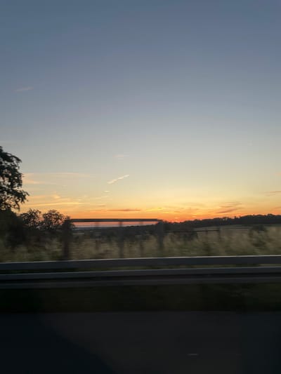 The image depicts a view from a moving vehicle, likely on a highway, overlooking a rural landscape at sunset. The sky is a gradient of soft blues and oranges, with wispy clouds and what appears to be a contrail from an aircraft. In the middle ground, a line of trees forms a silhouette against the horizon, with a strip of water or a large field visible behind them. A metal guardrail runs horizontally across the lower portion of the image, indicating the perspective from inside or alongside a road. The foreground is dominated by the dark asphalt of a road. There is no discernible location in the city of Essen, Germany, within the image. The scene is devoid of people or specific objects that would pinpoint a geographical location.