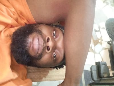 A close-up image, oriented 90 degrees clockwise, reveals an adult individual with a dark complexion and a full dark beard and mustache. The individual's face, visible from the chin to the forehead, features open eyes looking directly at the camera. The lips are full. An orange garment is visible on the left side of the frame, covering part of the upper body. A bare arm, consistent with the subject's skin tone, extends from the top right towards the bottom right. The background includes a horizontal wooden surface behind the subject's head. To the right and bottom, a tiled floor with light and dark geometric patterns is visible. A dark-colored object, appearing to be a stand or base for an electronic device, with a partial grill, rests on the tiled floor in the lower right. The scene is located in Magboro, Nigeria.