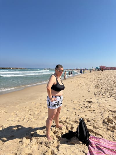 A medium shot captures a woman standing on a sandy beach in Ashkelon, Israel. The woman, wearing a black sports bra and black and white patterned shorts, faces the viewer with her left hand on her hip and her right hand slightly bent. She has short, dark hair, sunglasses, and a watch on her left wrist. A tattoo is visible on her left calf. Behind her, the Mediterranean Sea rolls with white-capped waves, and breakwaters extend into the water. The beach is populated with several people walking, and further in the background, there are beach umbrellas, tents, and structures, including a pink building. A black backpack and a striped beach towel are on the sand to the right of the woman. The sky above is a clear, bright blue.