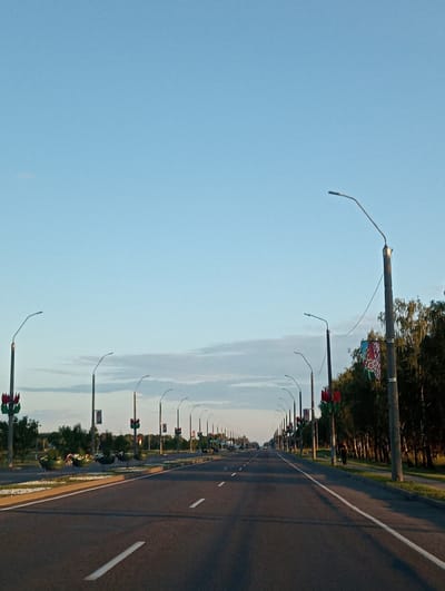 The image shows a long, paved road in a rural setting with streetlights lining the side. The road curves gently into the distance, with a white line marking the edge. Trees and some decorative elements, possibly flags or banners, are visible behind the streetlights. The sky is a clear, pale blue, suggesting it is daytime, likely morning or late afternoon due to the soft lighting. The overall mood is peaceful and serene. There is no visible text in the image.